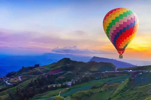 Colorful hot air balloon flying over lush green mountains during sunset